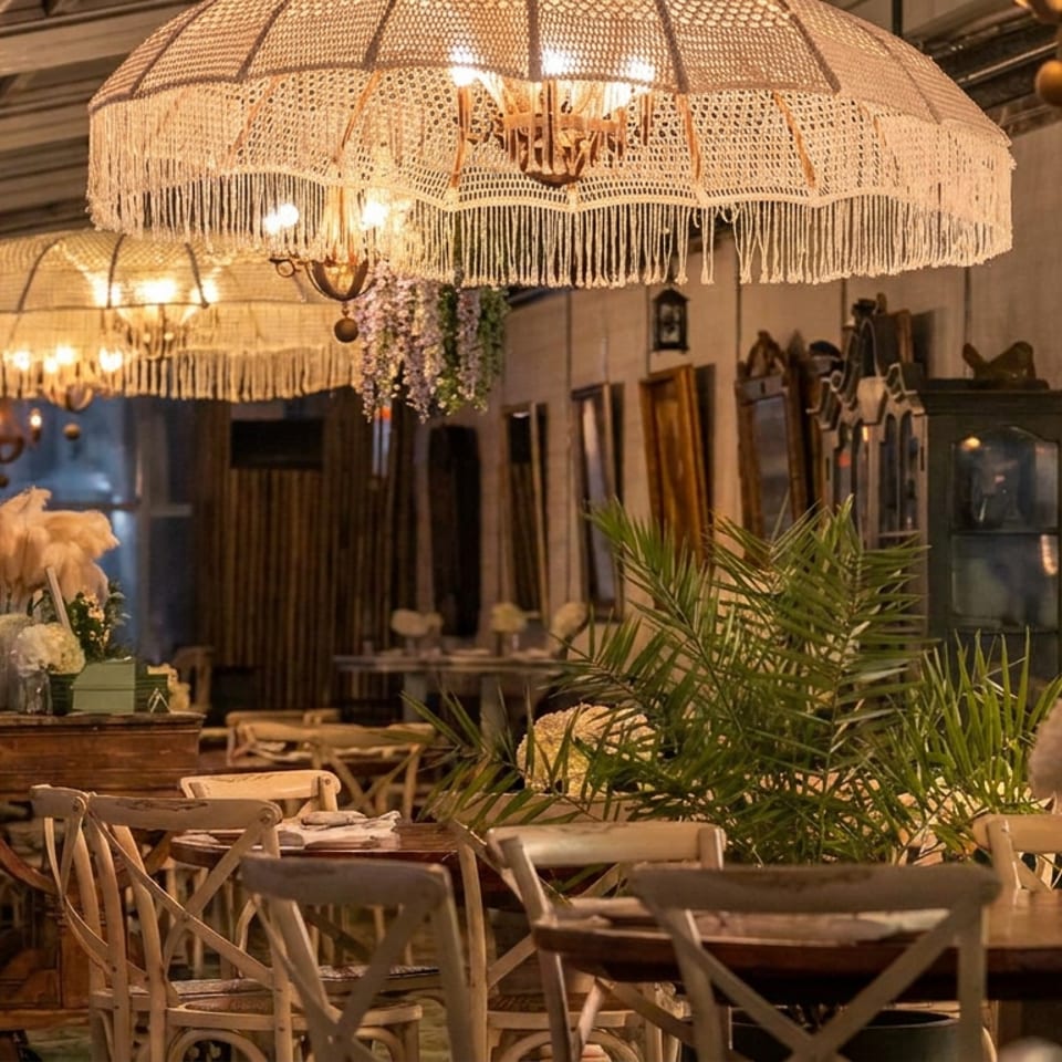 Warmly lit restaurant interior with fringed chandeliers, wooden tables, vintage furnishings, and lush tropical plants.