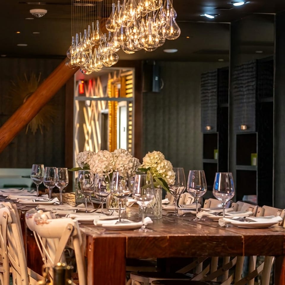Elegant restaurant dining room with a long set table, wine glasses, floral centerpieces, wood accents, and hanging glass chandeliers.