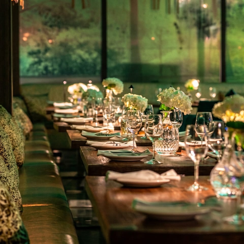 Elegant restaurant dining room with upholstered banquette seating, patterned pillows, set tables, flowers, and large garden-view windows.