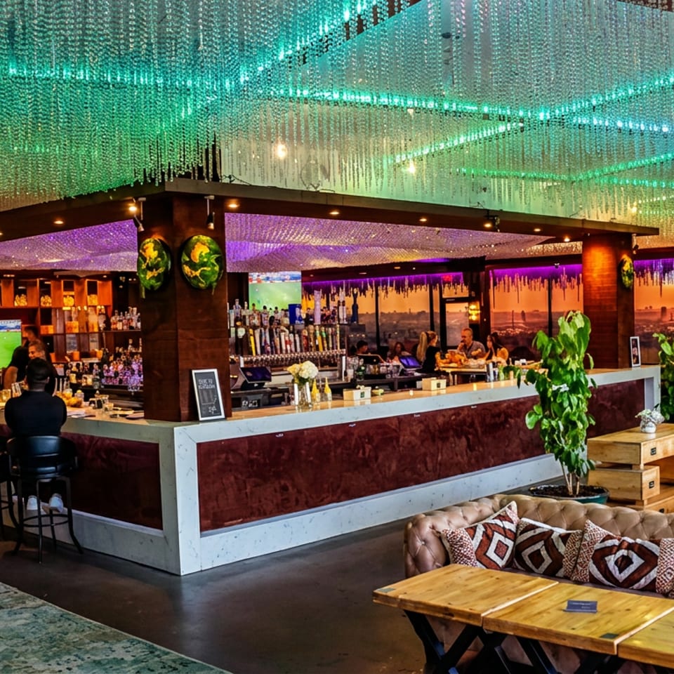 Vibrant sports bar interior with glowing beaded ceiling, central bar, televisions showing soccer, dining tables, and city views.