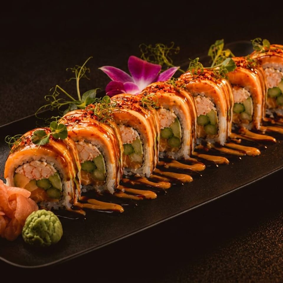 Sushi roll topped with orange sauce and garnishes, served on a black plate with pickled ginger and wasabi.