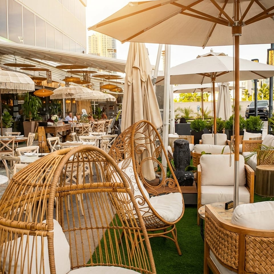 Outdoor restaurant patio with wicker seating, white umbrellas, set dining tables, greenery, and city buildings in the background.